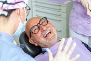An older man with glasses smiles while reclining in a dental chair, happily interacting with a masked dental professional and another staff member in a clinic. Dental equipment and cabinets appear in the background.
