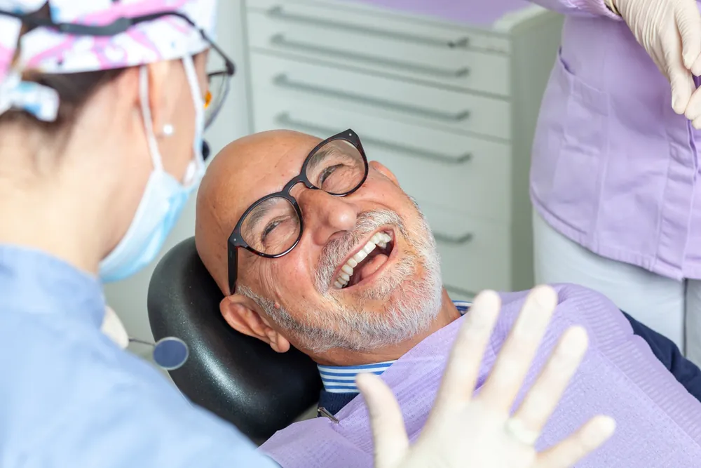 An older man with glasses smiles while reclining in a dental chair, happily interacting with a masked dental professional and another staff member in a clinic. Dental equipment and cabinets appear in the background.