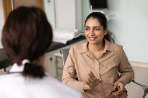A woman in a tan shirt smiles and gestures while speaking with another person, possibly a doctor, in a medical exam room. The focus is on the smiling woman while the other person is visible from behind.