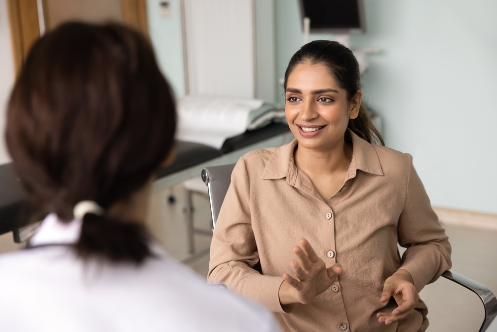 A woman in a tan shirt smiles and gestures while speaking with another person, possibly a doctor, in a medical exam room. The focus is on the smiling woman while the other person is visible from behind.