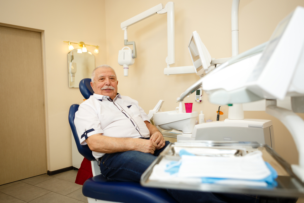An older man with a mustache sits in a dental chair, smiling. Dental equipment, tools on a tray, and a pink cup are visible nearby. The room is clean and brightly lit with beige walls.